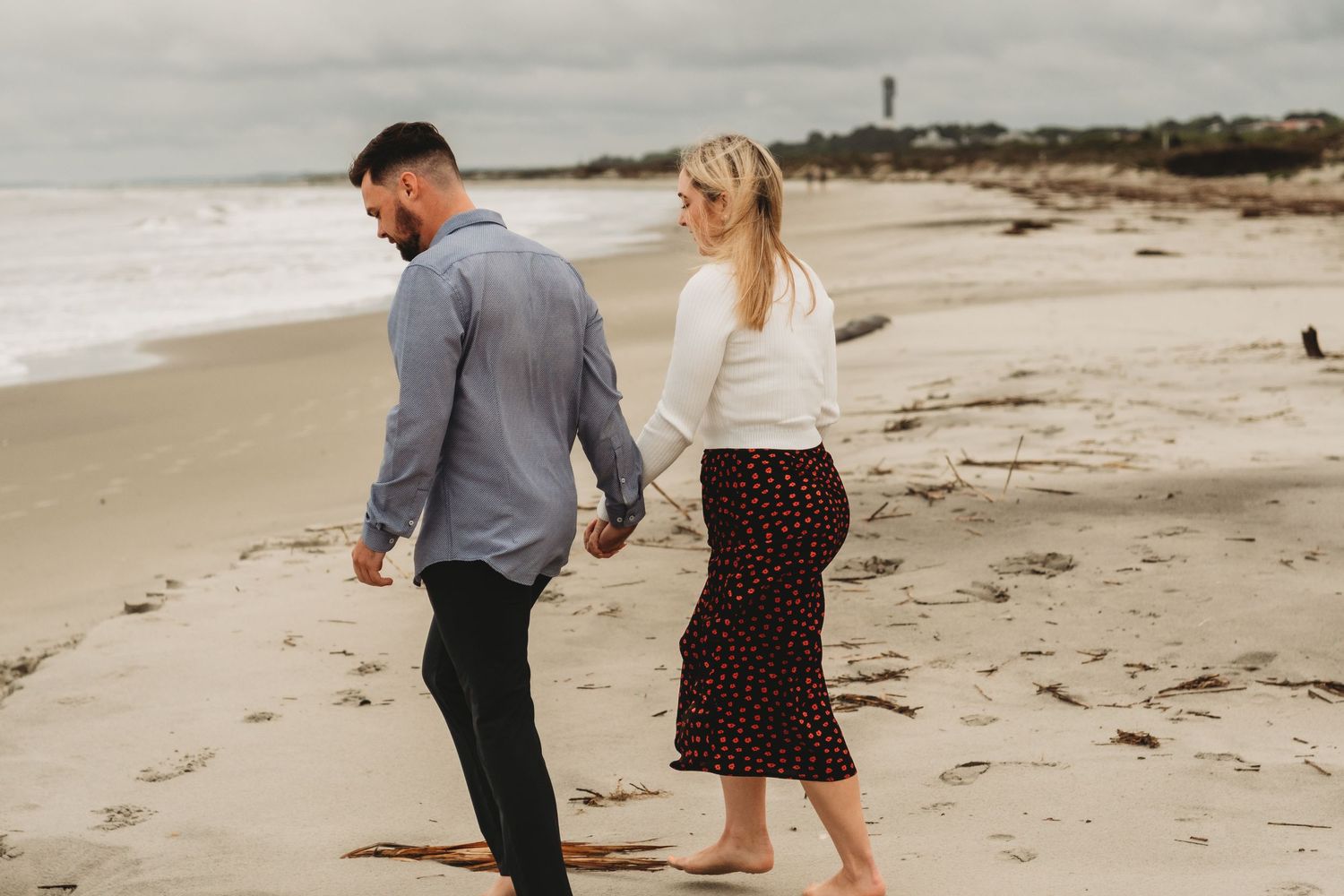 Couple holding hands walking barefoot along sandy beach on an overcast day.