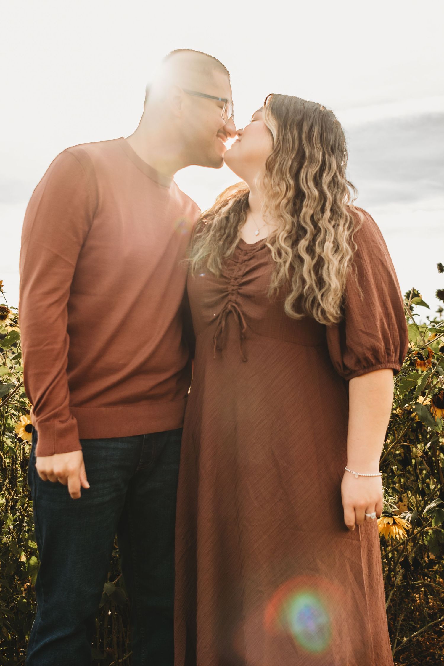 A couple nearly kissing in a sunflower field at golden hour, both wearing warm brown tones.