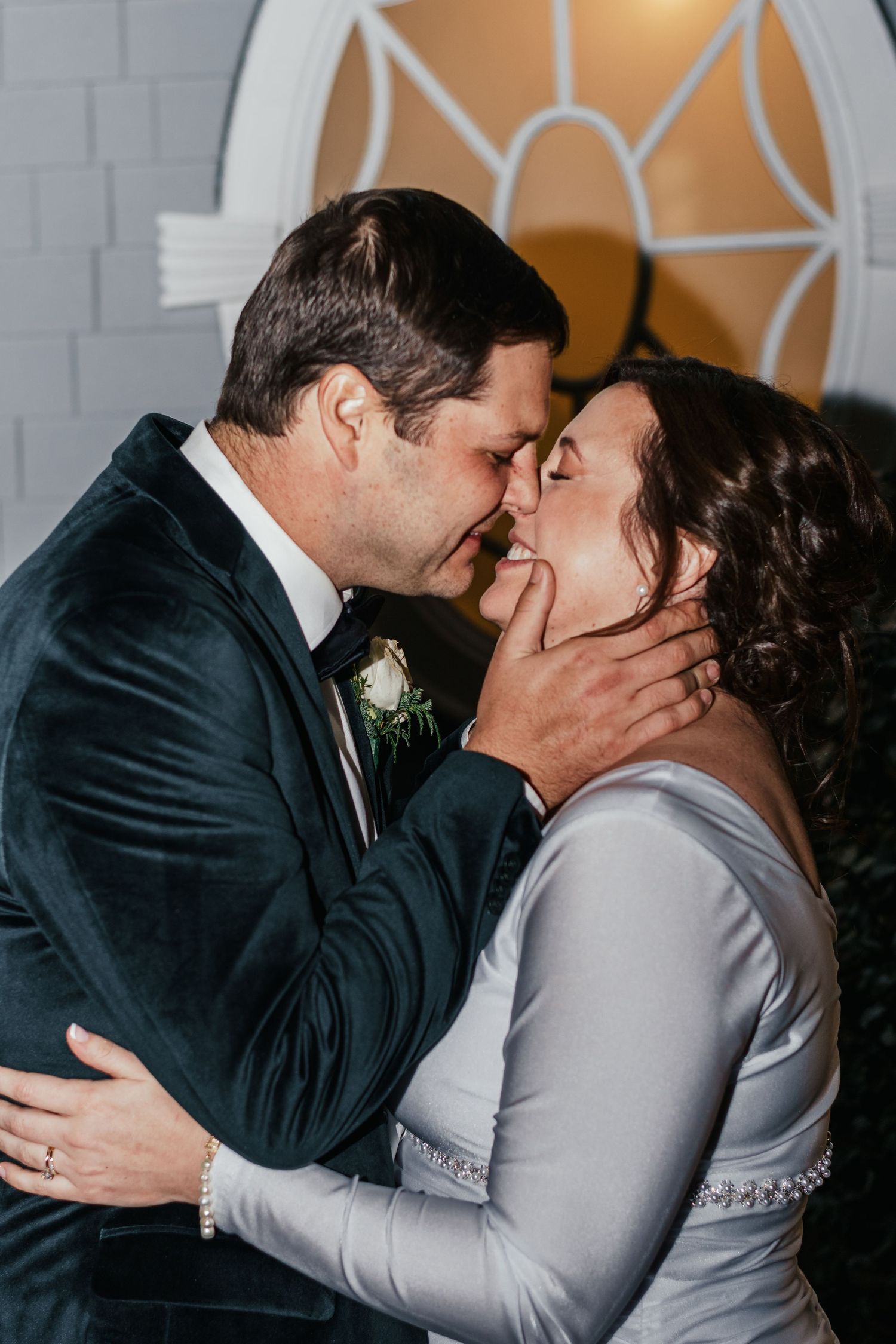 Bride and groom share passionate first kiss at wedding ceremony near decorative window.