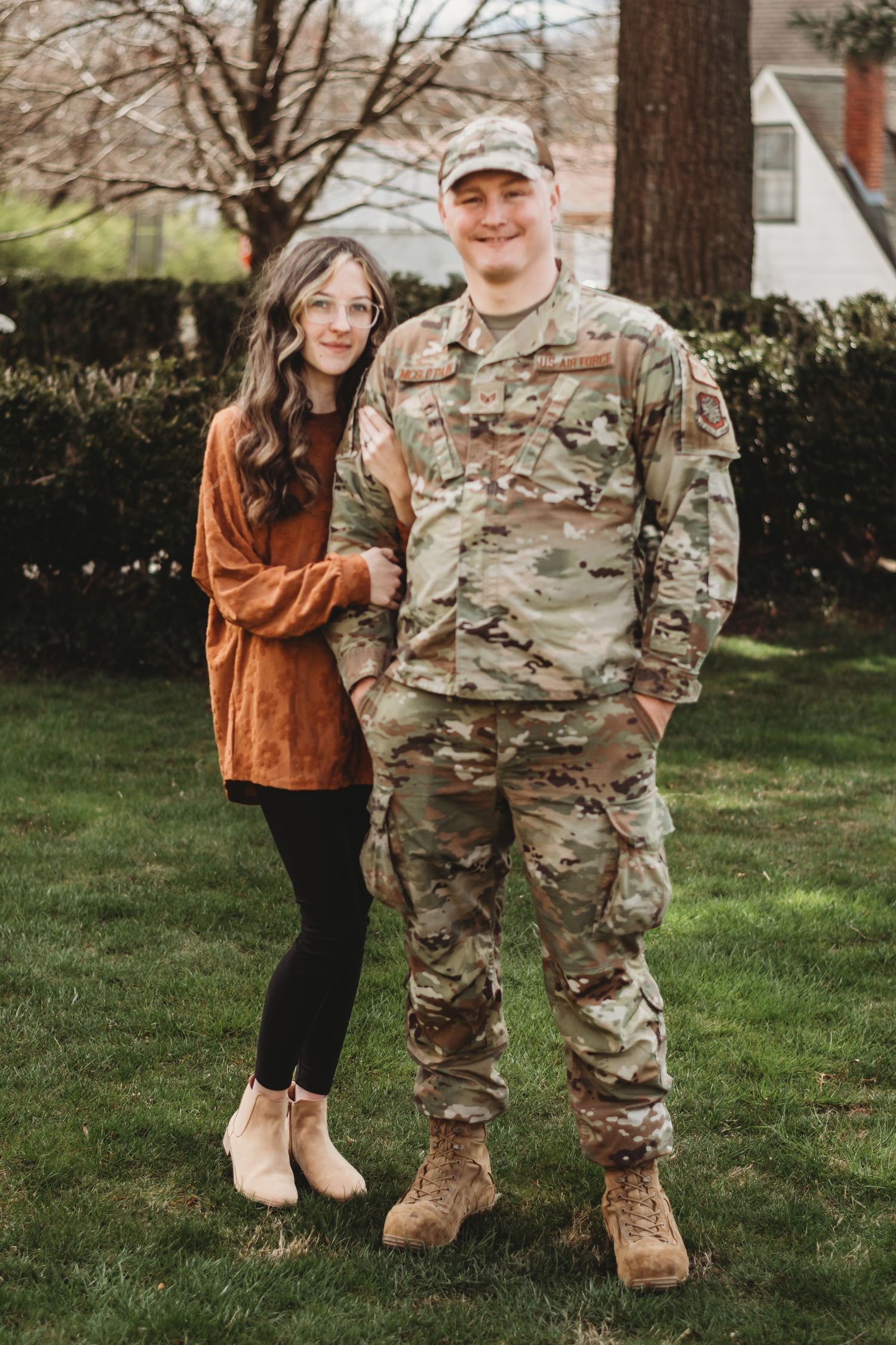 A soldier in camouflage uniform stands proudly with a woman in a rust-colored cardigan outdoors.