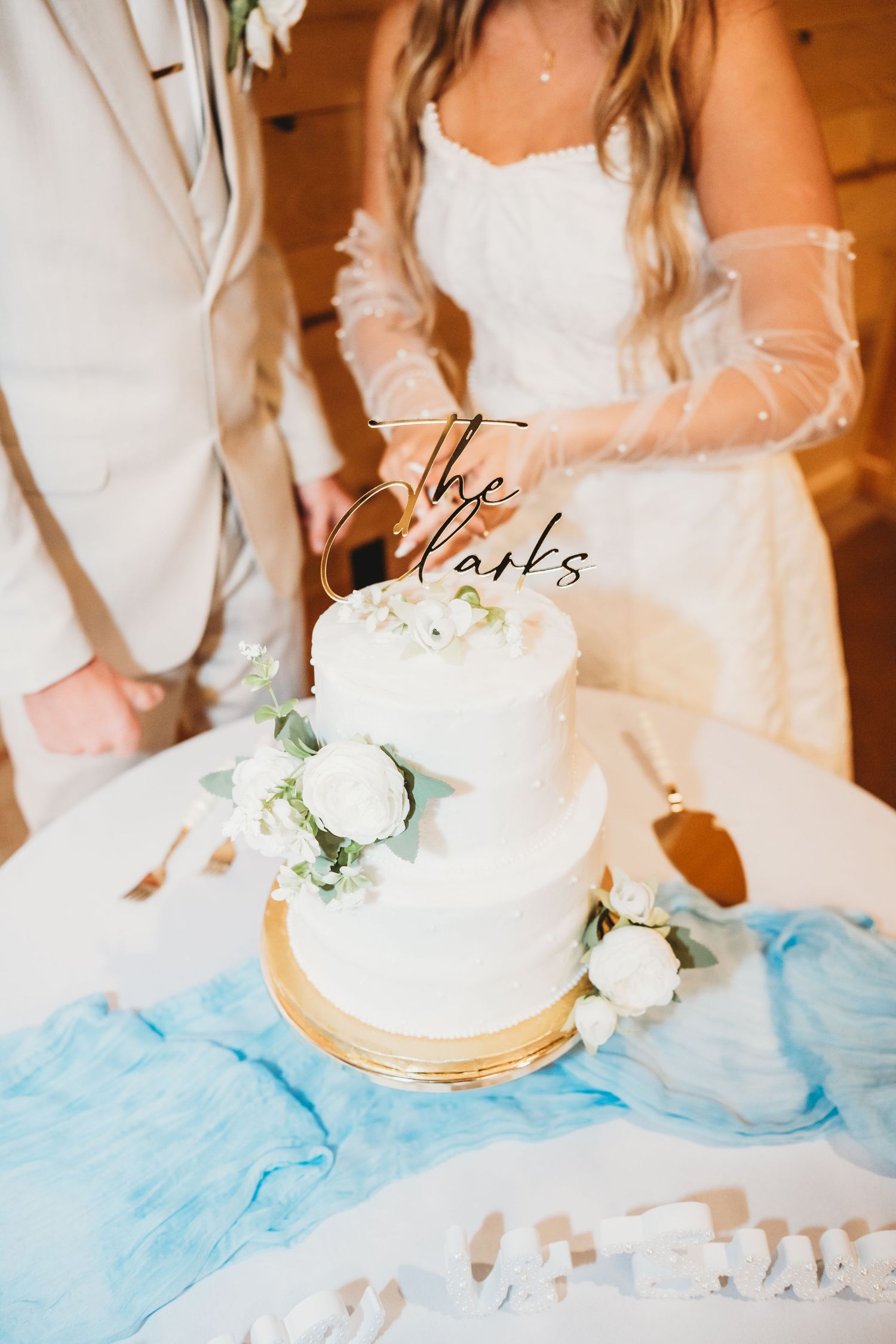 Two-tier white wedding cake with roses and The Clarks topper displayed at wedding reception.