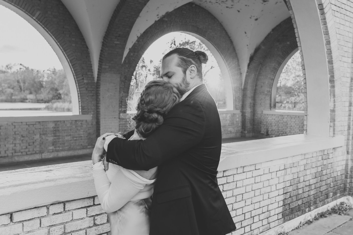 Black and white photo of couple embracing under arched brick bridge.