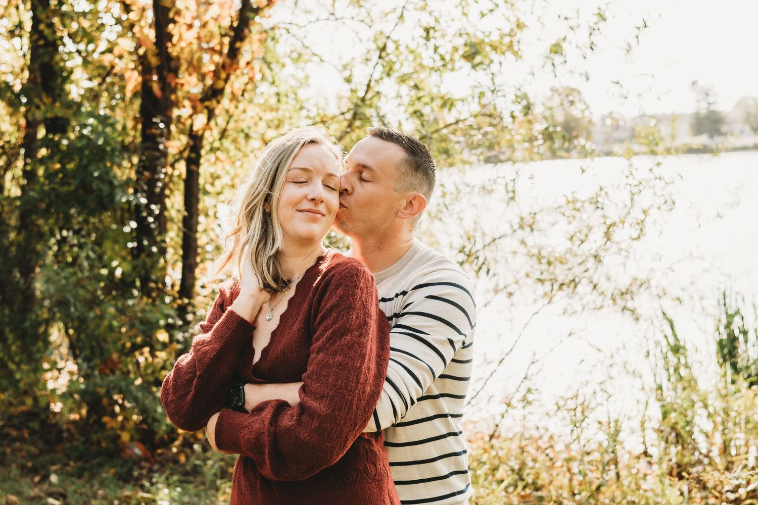 A couple embracing near a sunlit lake in autumn, man kissing woman's cheek from behind.