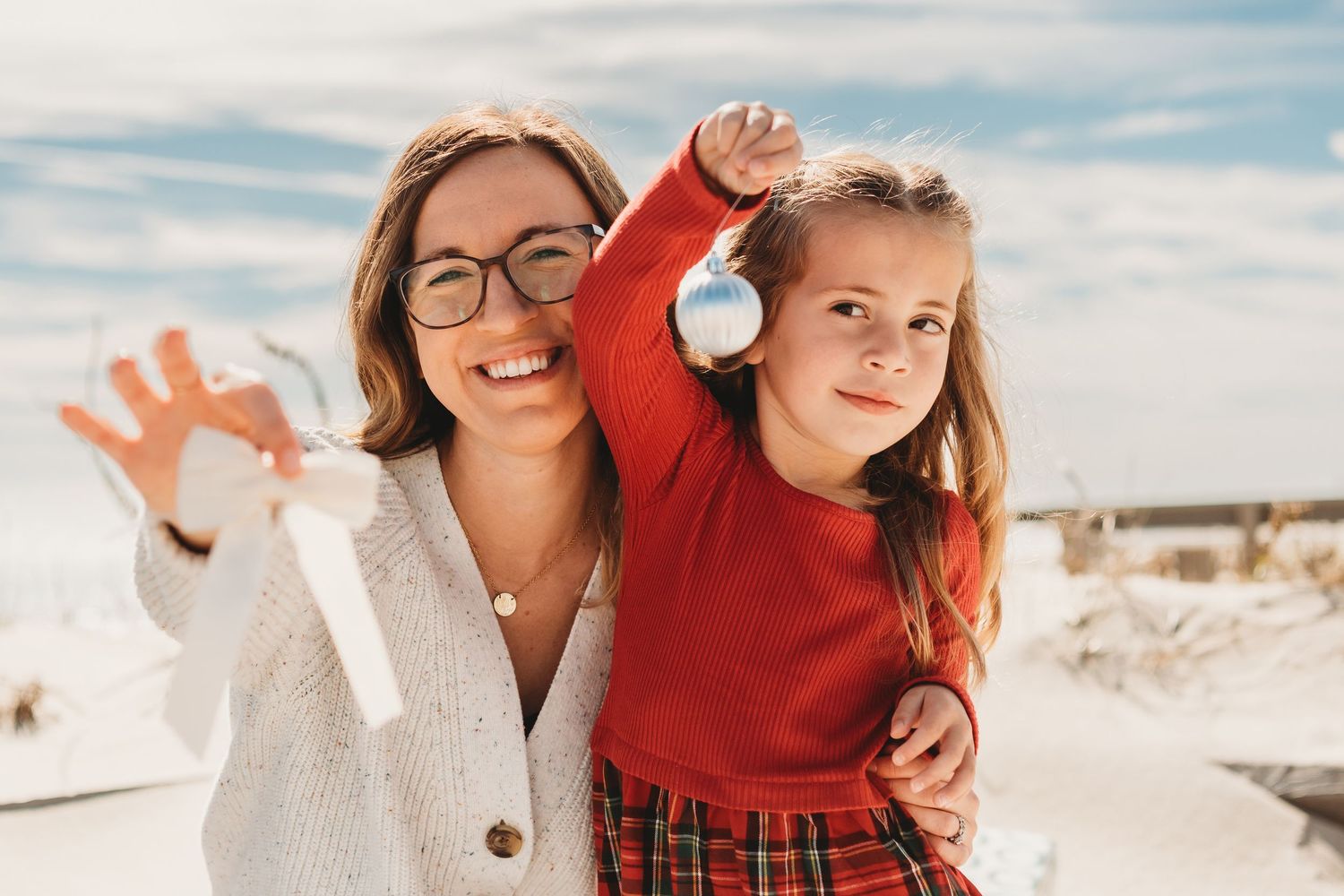 A smiling woman with glasses and a young girl hold Christmas ornaments outdoors on a sunny day.