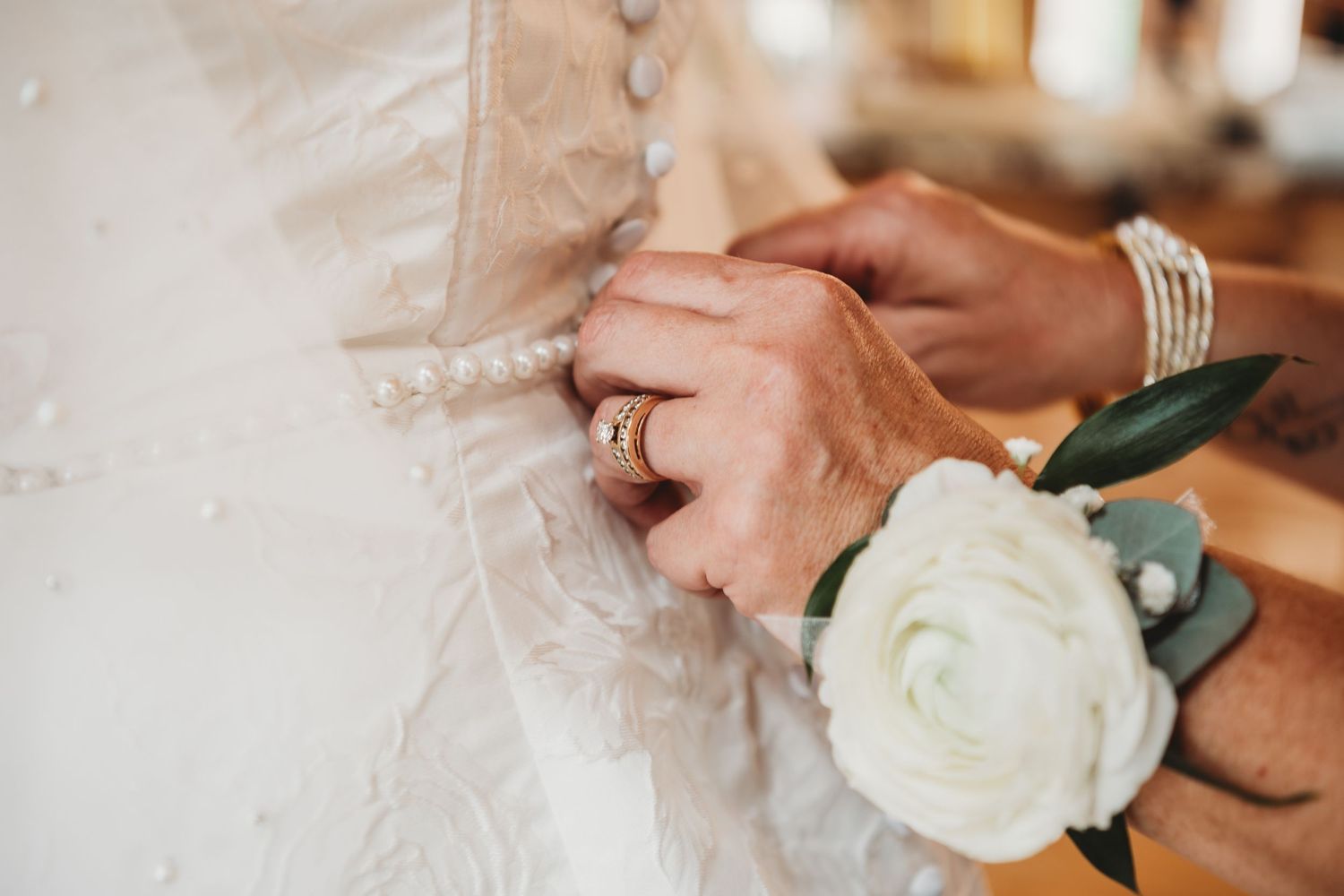 Close-up of hands with corsage buttoning the back of a pearl-detailed wedding dress.