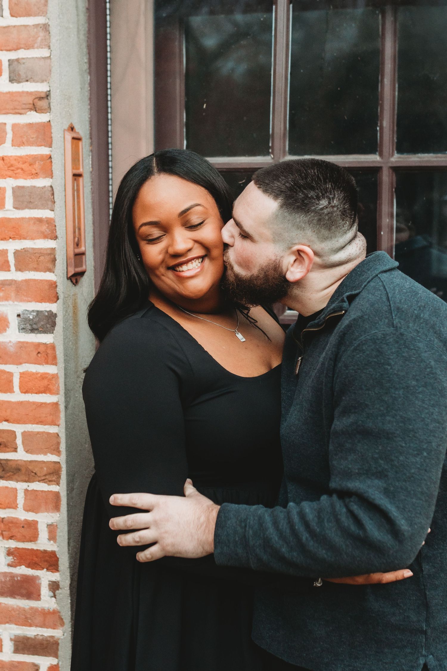 A man kisses a smiling woman on the cheek as they embrace near a brick wall and window.