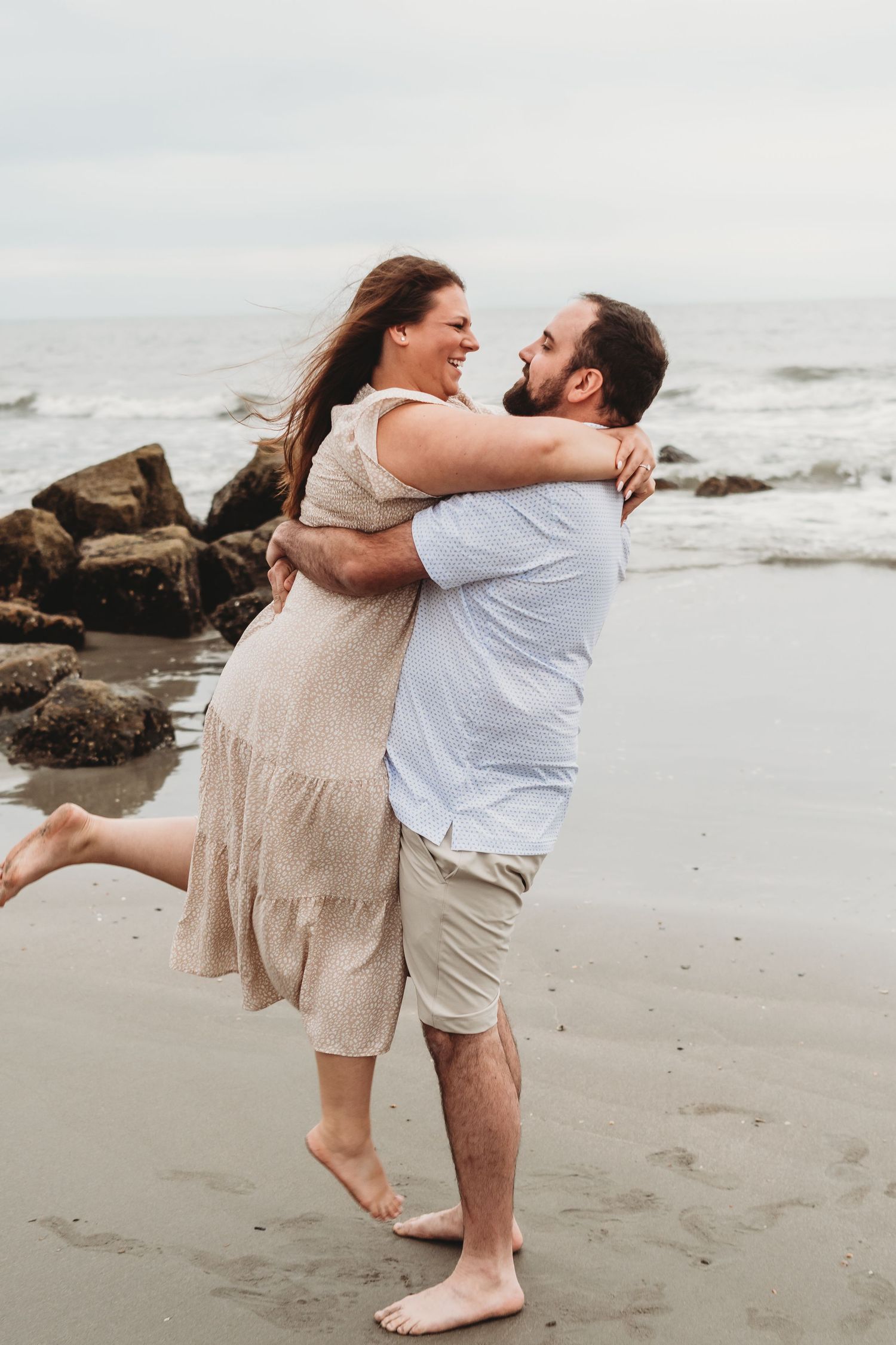 Laughing couple embracing joyfully on a rocky beach shoreline with ocean waves in the background.