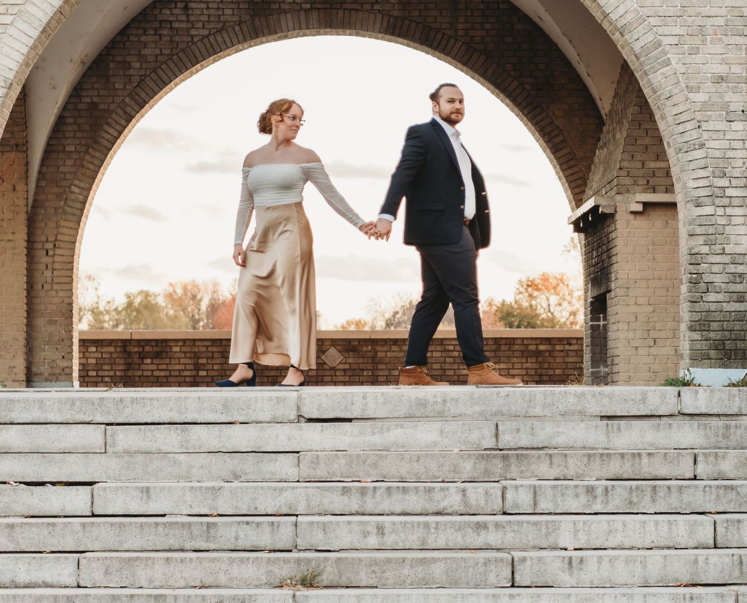 Couple holding hands walking down stone steps under a brick arch at sunset.