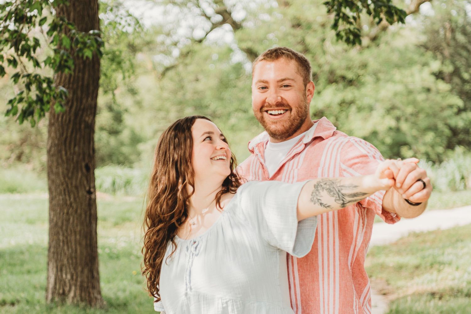 A joyful couple dancing outdoors in a park, man in pink shirt and woman in white off-shoulder dress.