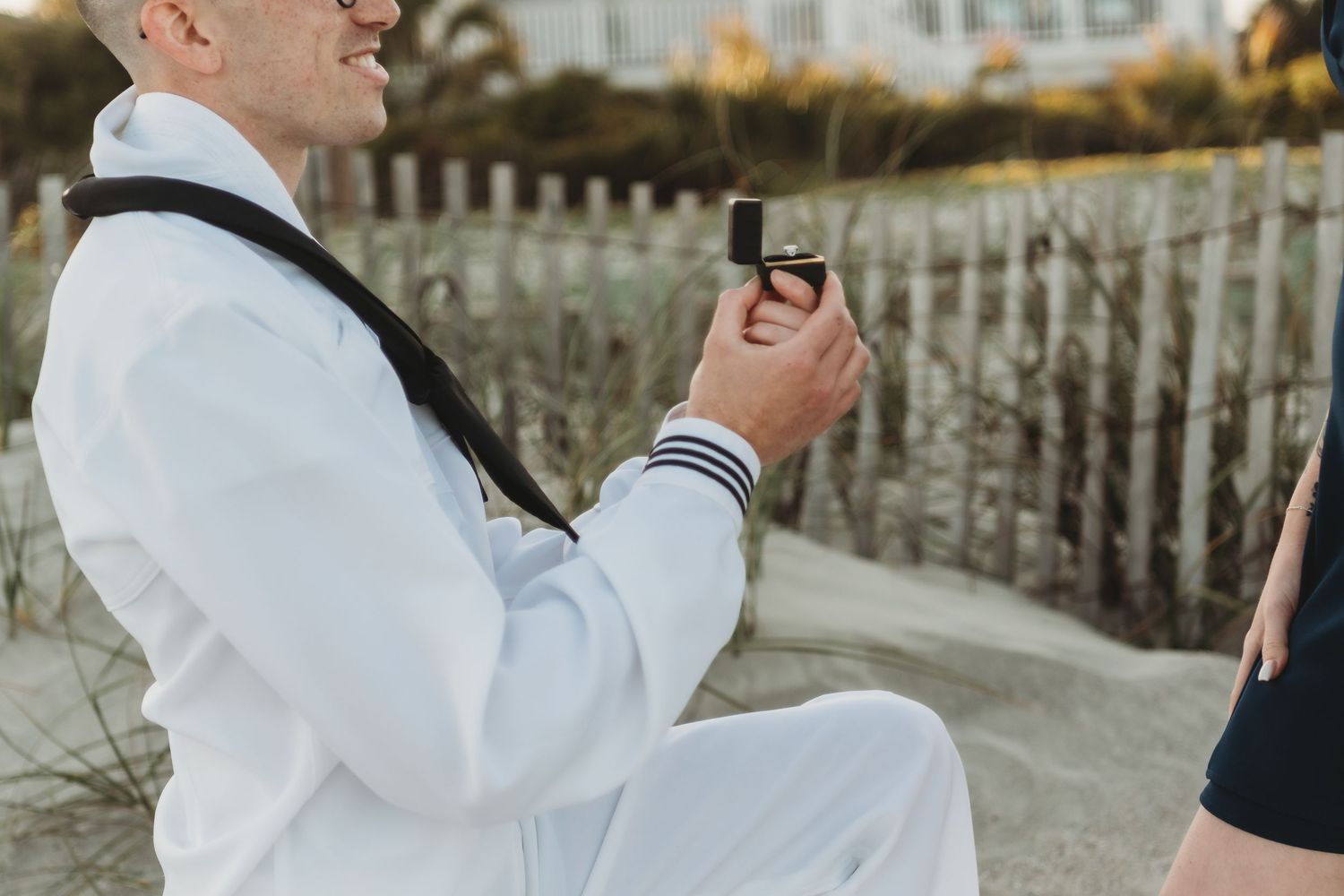 Man in white suit kneeling on sandy beach holding a ring box during a romantic marriage proposal.