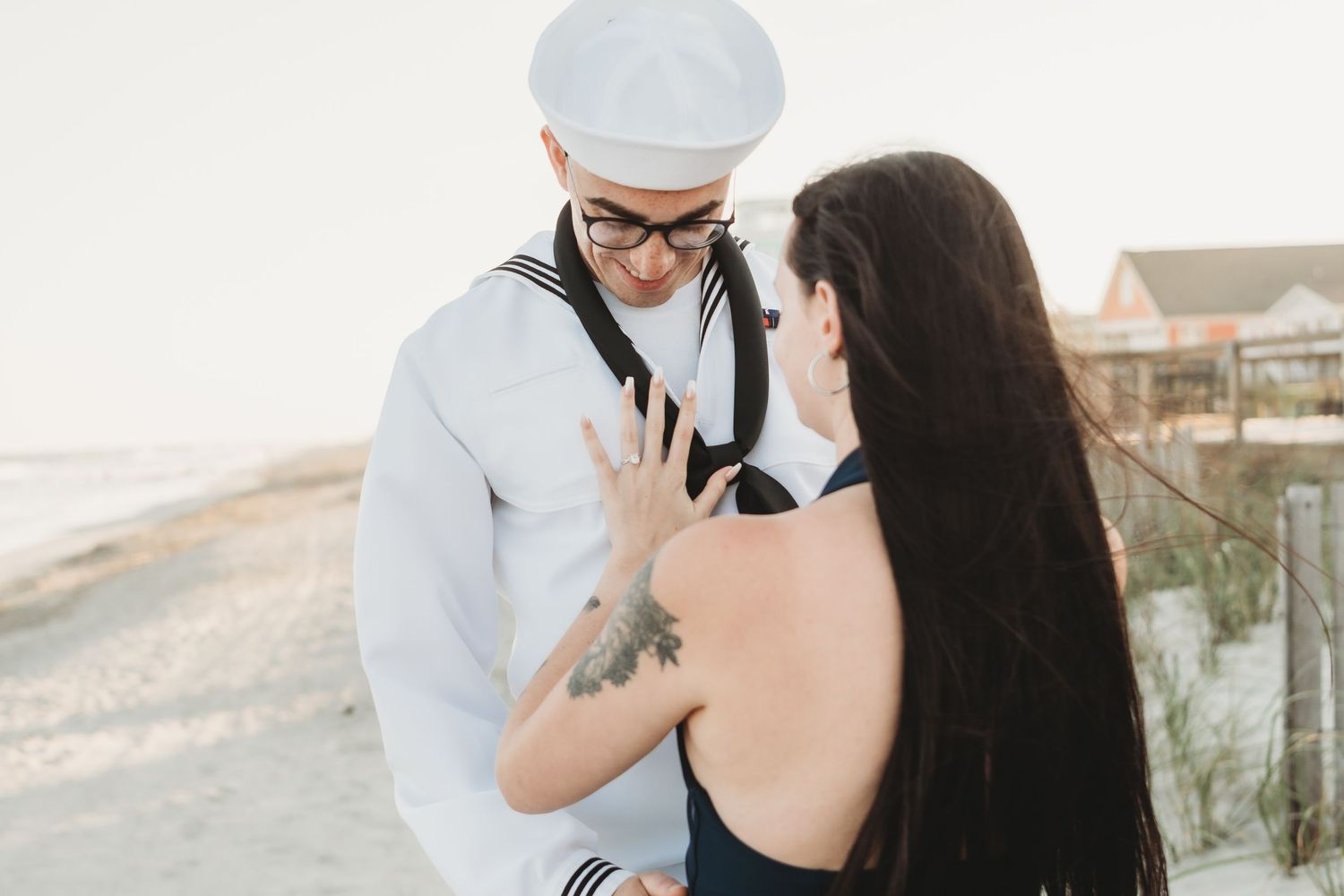 A sailor in white uniform gazes lovingly at a dark-haired woman on a sandy beach at sunset.