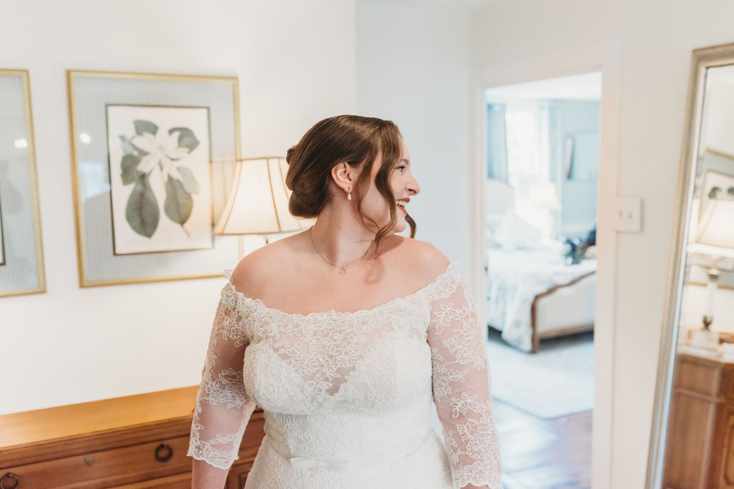 Bride in off-shoulder lace wedding gown with long sleeves standing in a softly lit room.