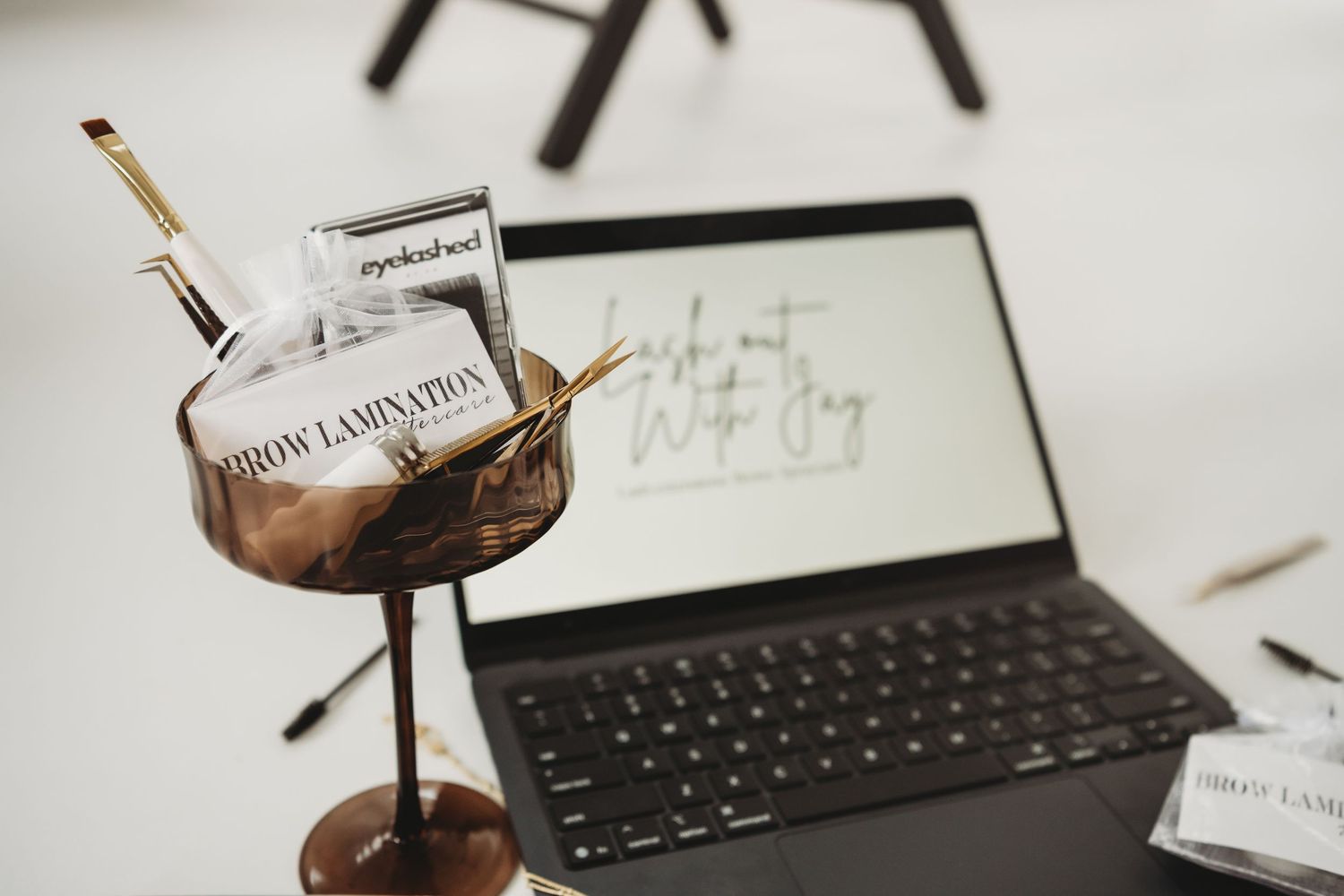 Brow lamination beauty tools displayed in a decorative bowl beside an open laptop on a white desk.
