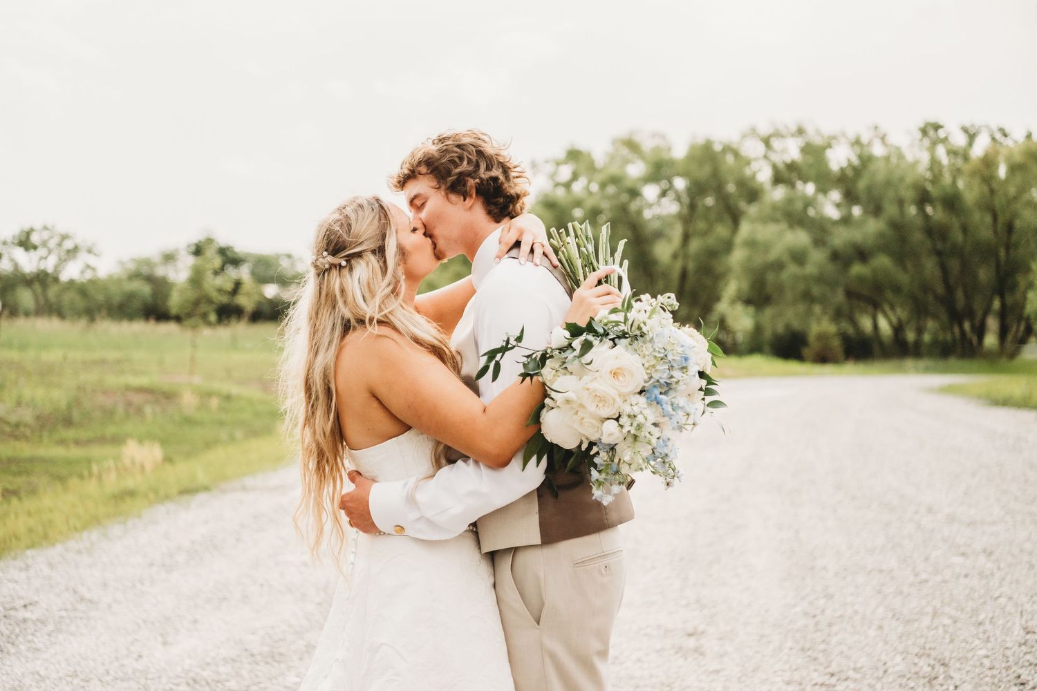 Bride and groom sharing a romantic kiss on a gravel road surrounded by green trees.