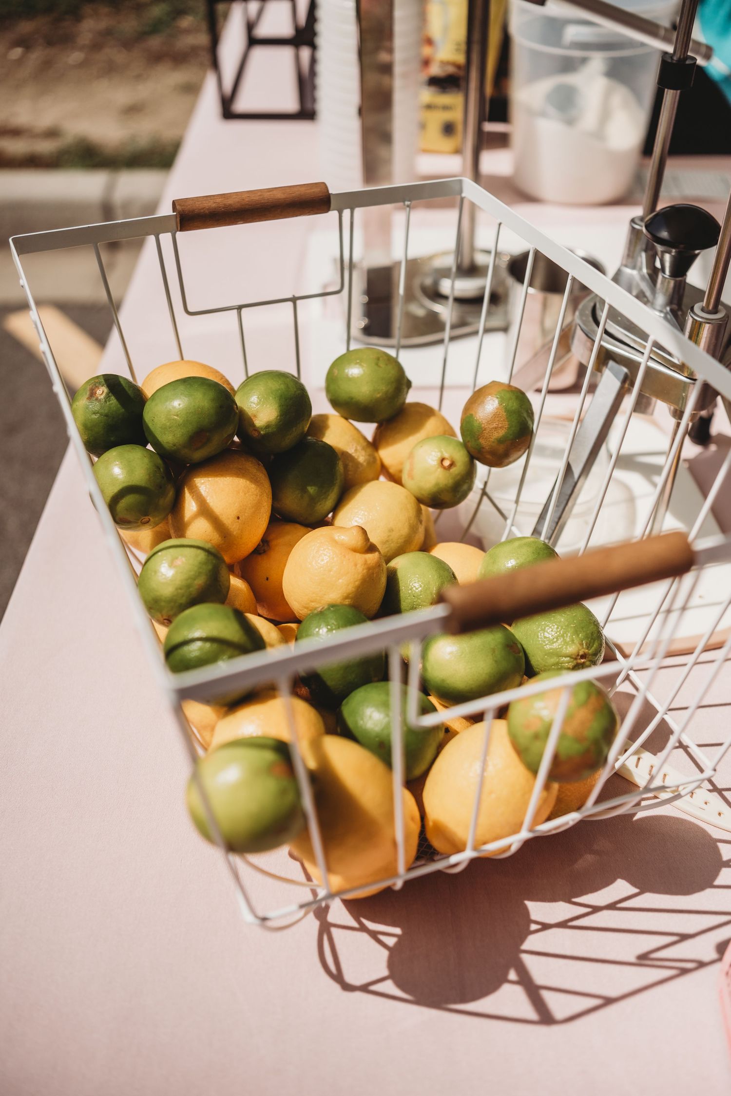 Wire basket filled with fresh lemons and limes on a bright sunny outdoor table surface.