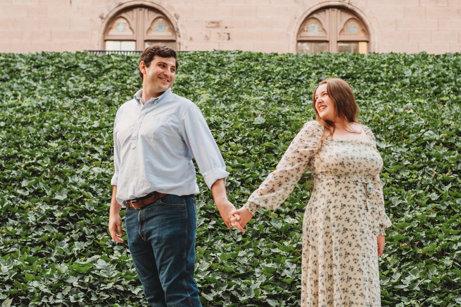 A smiling couple holds hands while walking in front of a lush green ivy-covered wall.