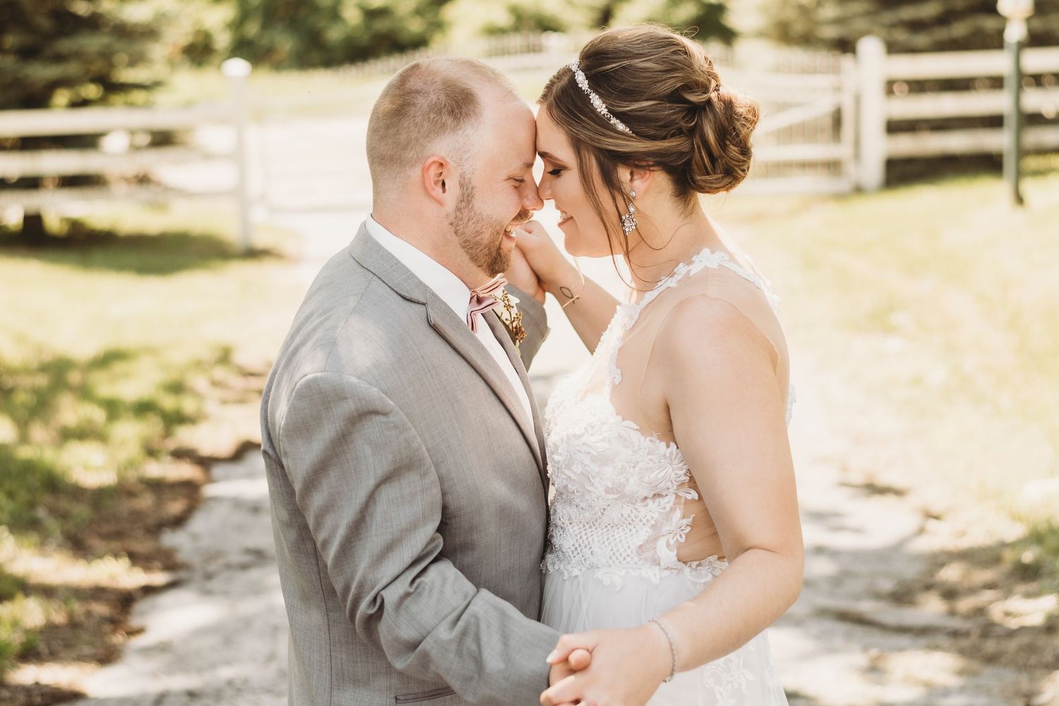 Bride and groom touching foreheads tenderly outdoors on a sunny wedding day near a wooden fence.