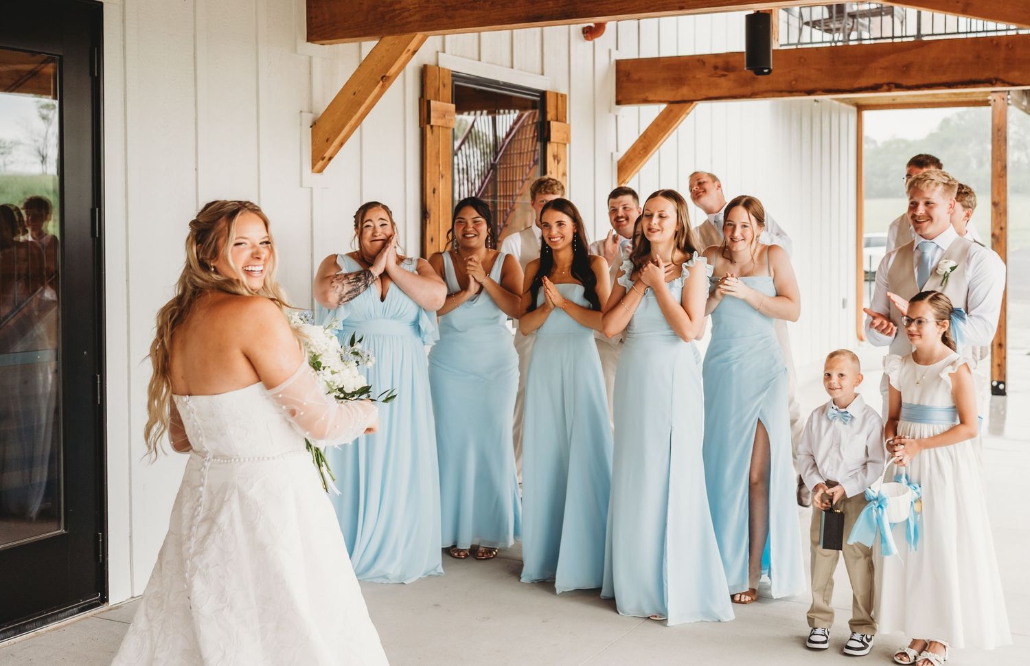 Bride revealing her wedding dress to bridesmaids in light blue gowns who react with excitement.