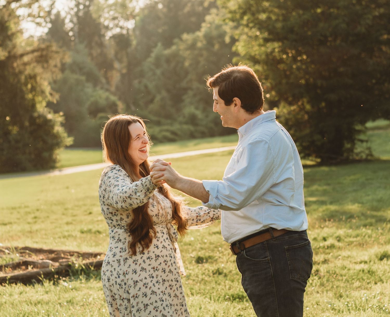A happy couple holds hands and laughs together in a sunlit grassy field at golden hour.