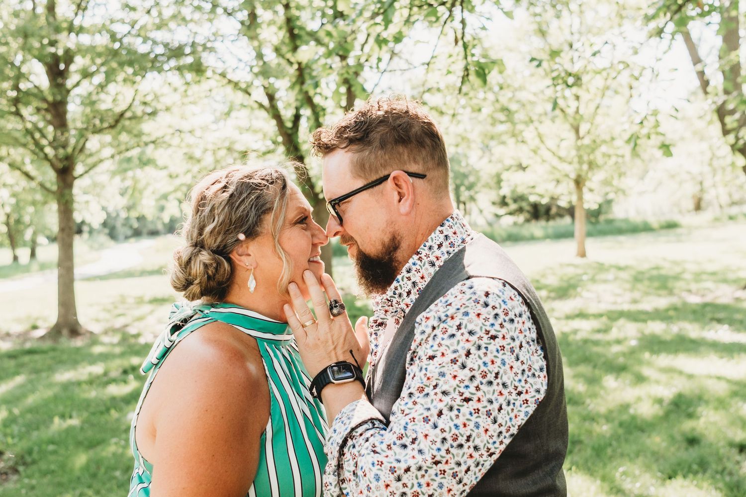 Couple nearly kissing in a lush green park, groom in floral shirt and vest, bride in teal dress.