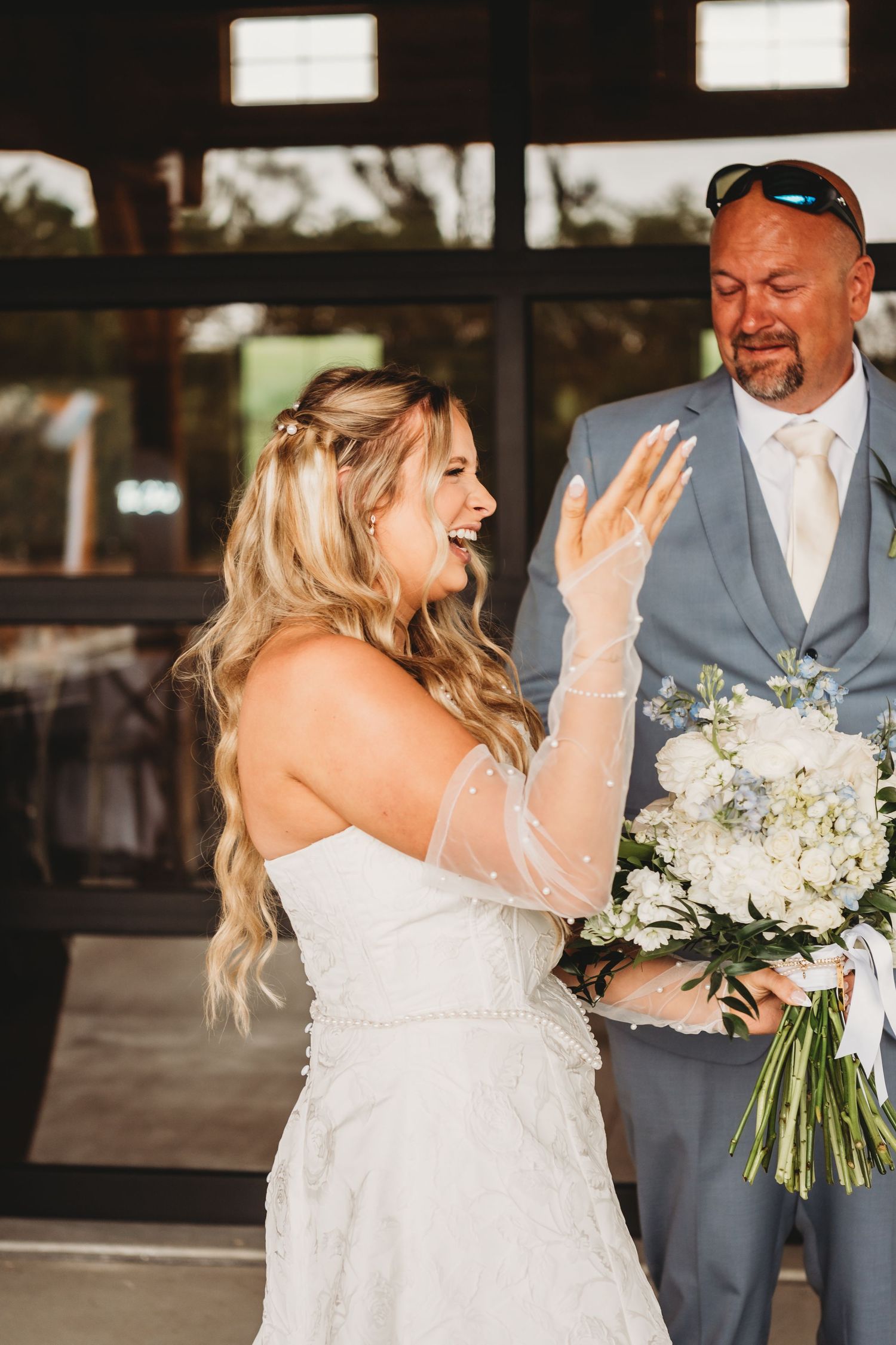 Excited bride in white gown showing her ring to a man in a gray suit holding her bouquet.