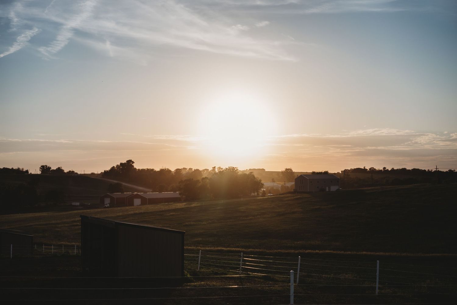 Golden sunset over a peaceful rural farm landscape with rolling green fields and silhouetted trees.