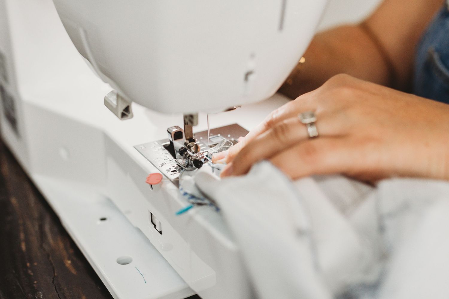 Close-up of hands using a white sewing machine to stitch white fabric, with a ring visible on one finger.
