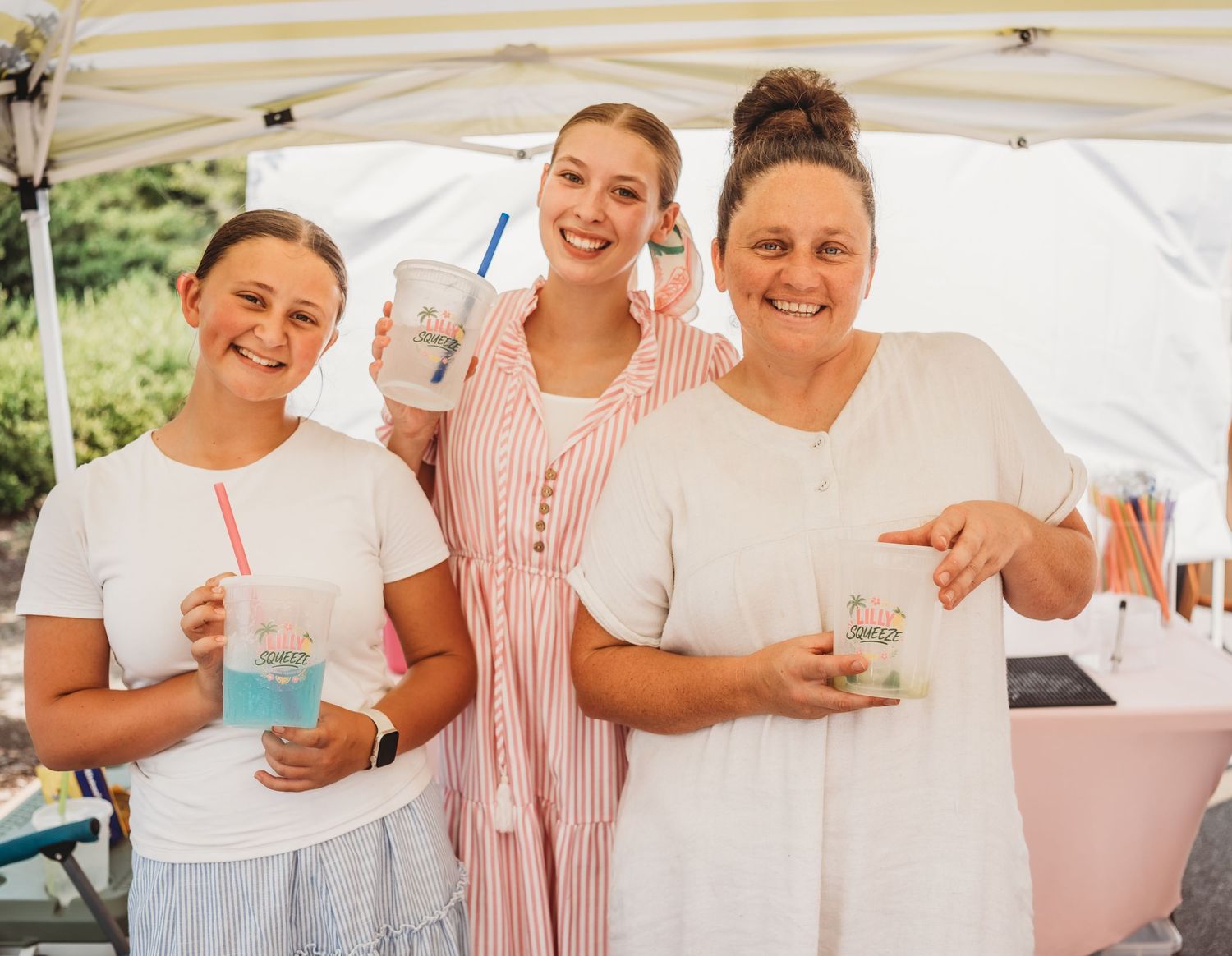 Three women smile holding colorful drinks at an outdoor summer event under a white canopy tent.