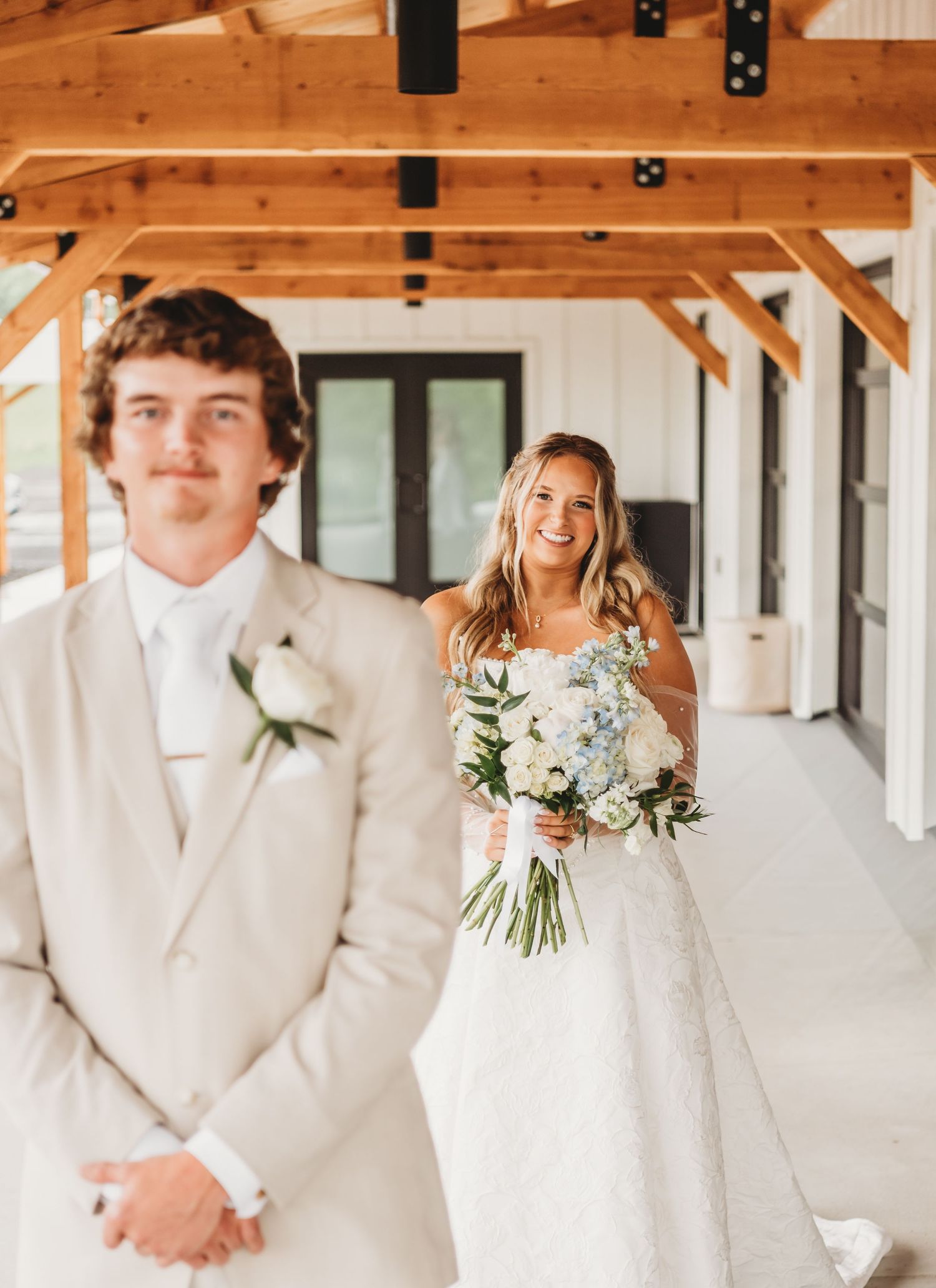 Bride smiling at groom during first look at a barn venue, holding blue and white floral bouquet.