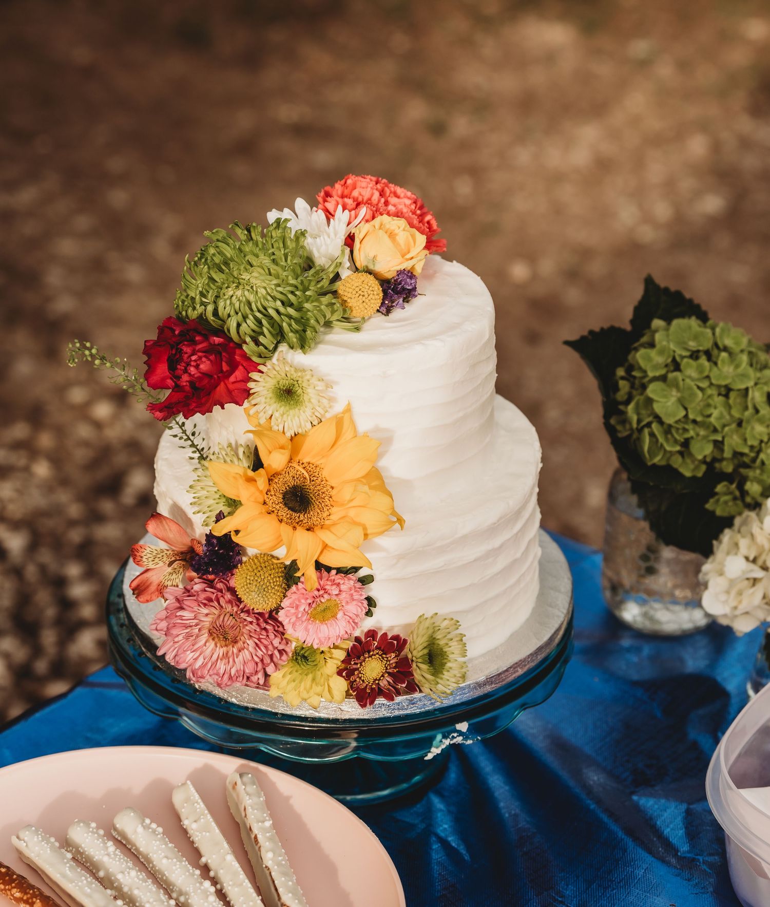 Two-tier white wedding cake decorated with colorful fresh flowers on glass stand.