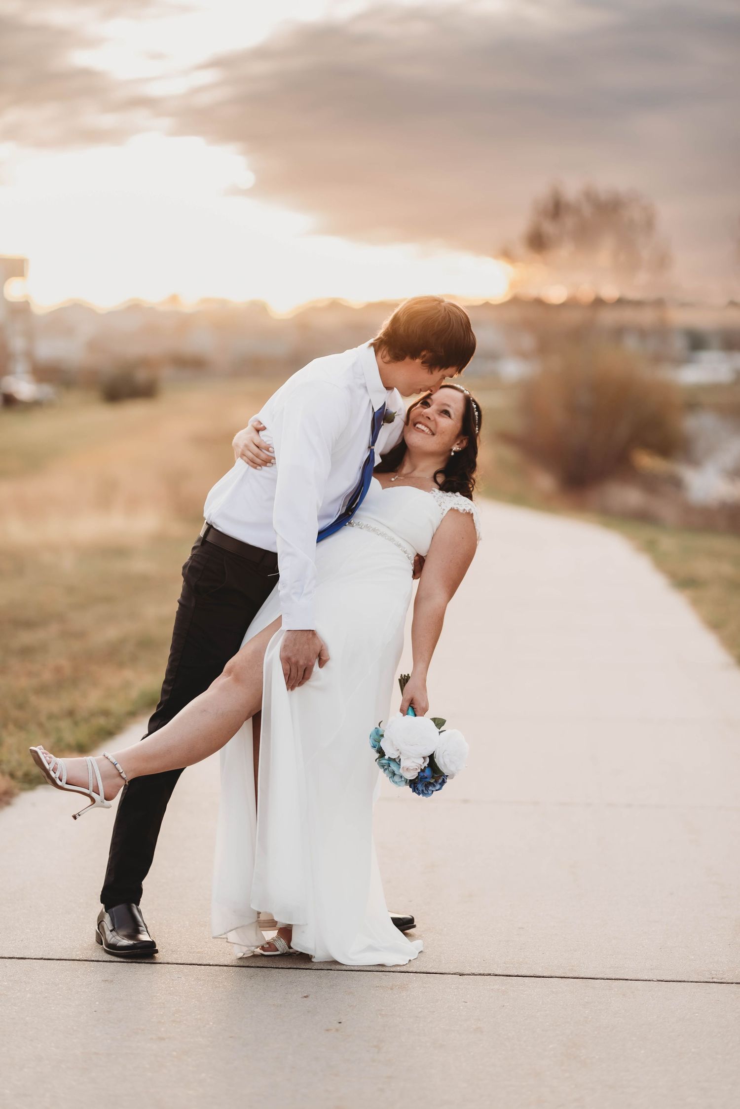 Groom dips bride in white dress holding blue bouquet at sunset outdoor wedding.