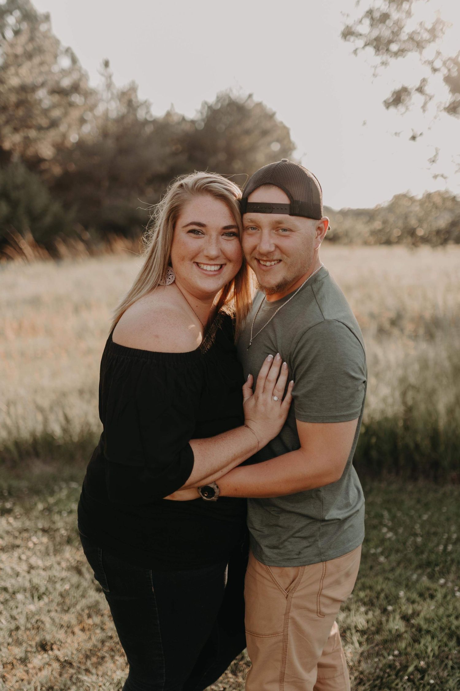 Smiling couple embracing in golden sunlit field, woman in black off-shoulder top.