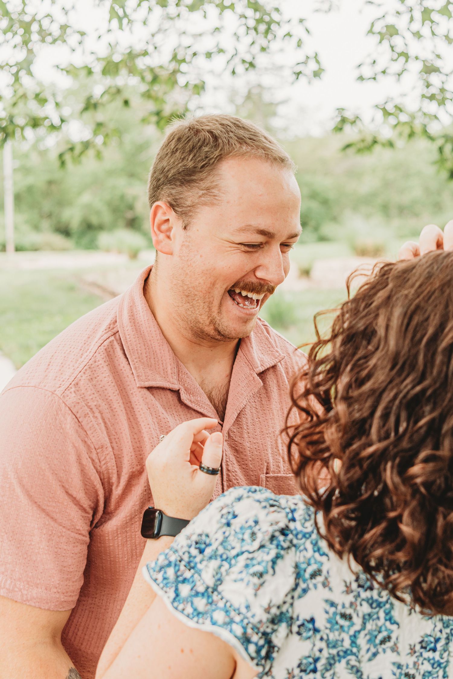 A laughing man in a pink shirt sharing a joyful moment with his curly-haired partner outdoors.