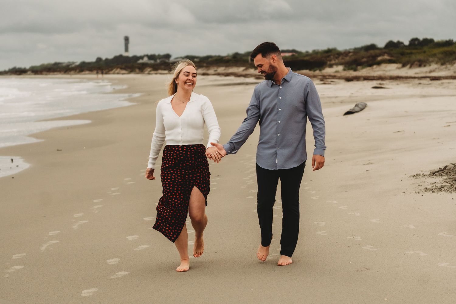 Couple walking hand in hand on beach with lighthouse visible in background.
