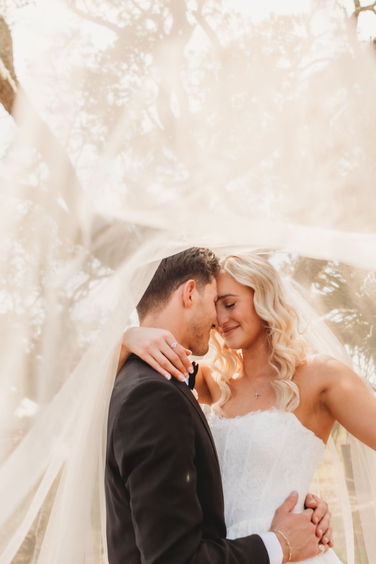 Bride and groom embracing tenderly under flowing veil in golden outdoor wedding portrait.