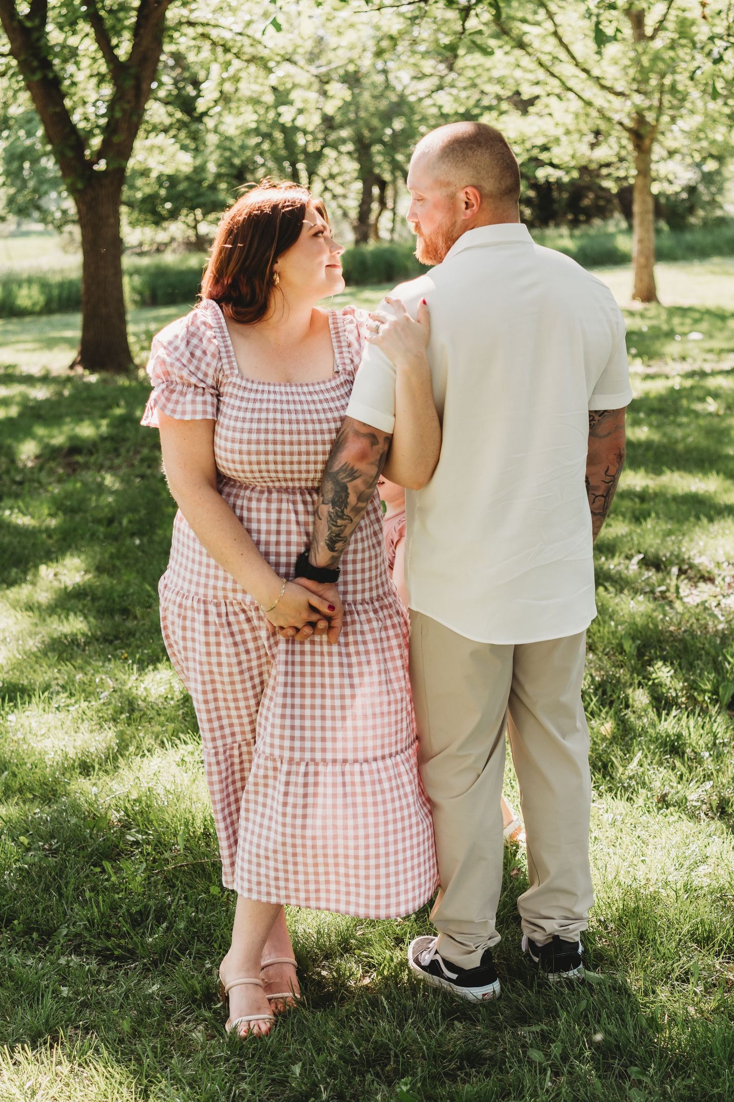 A tattooed couple walks hand in hand through a sunny green park, gazing at each other.