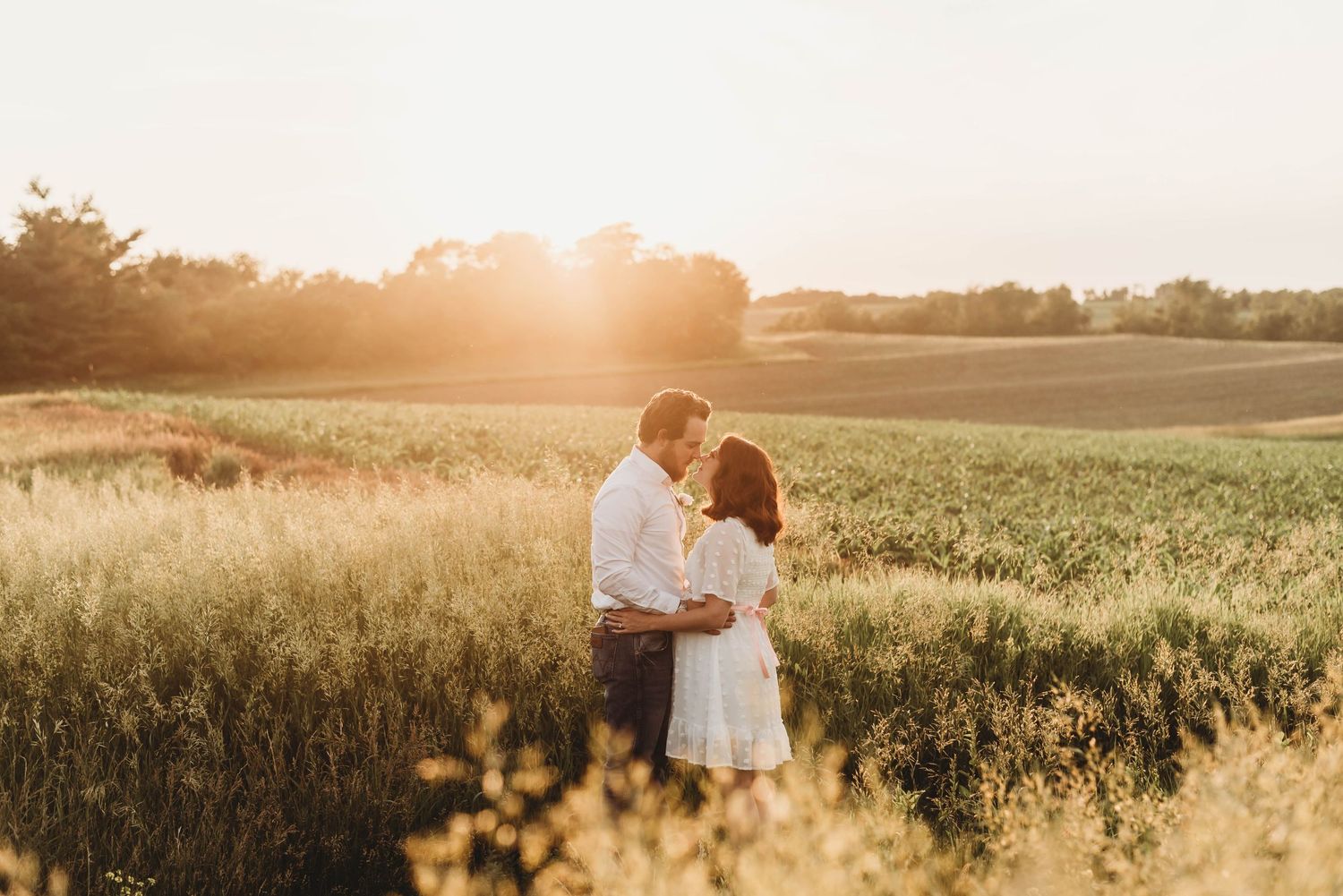 Couple embracing in golden sunset light in lush green field during romantic engagement session.