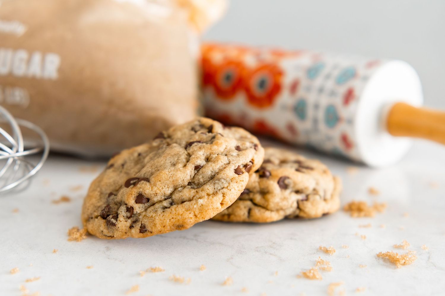 Freshly baked chocolate chip cookies on marble surface with brown sugar bag and rolling pin in background.