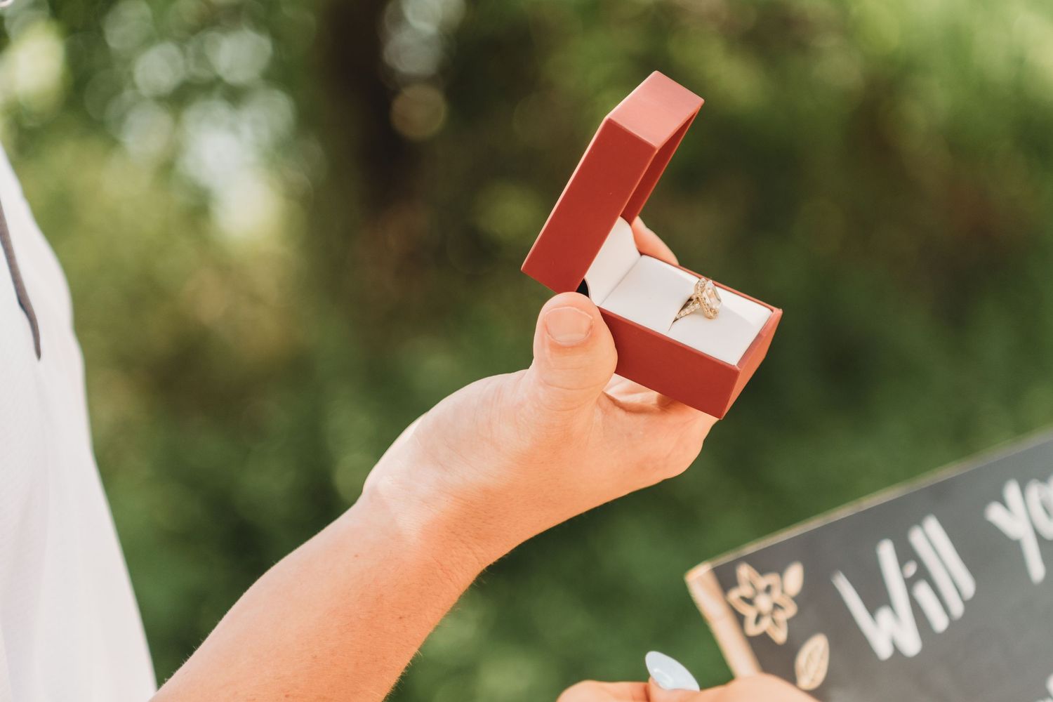 Hand holding open red ring box with diamond engagement ring during an outdoor marriage proposal.
