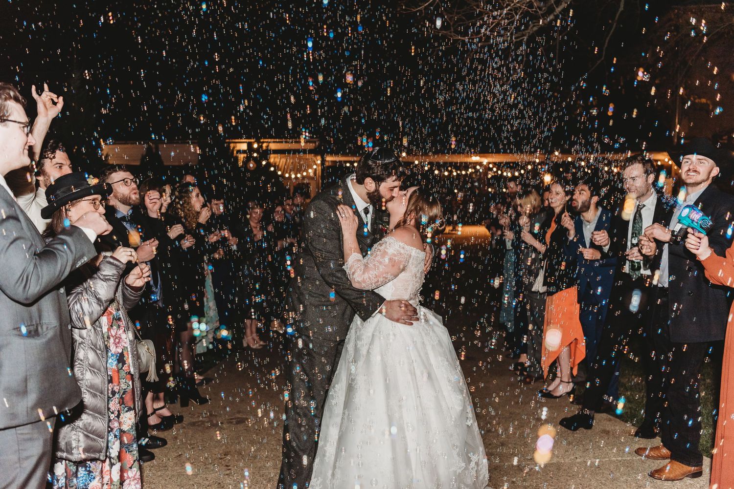 Bride and groom sharing a kiss surrounded by confetti and cheering wedding guests at night.