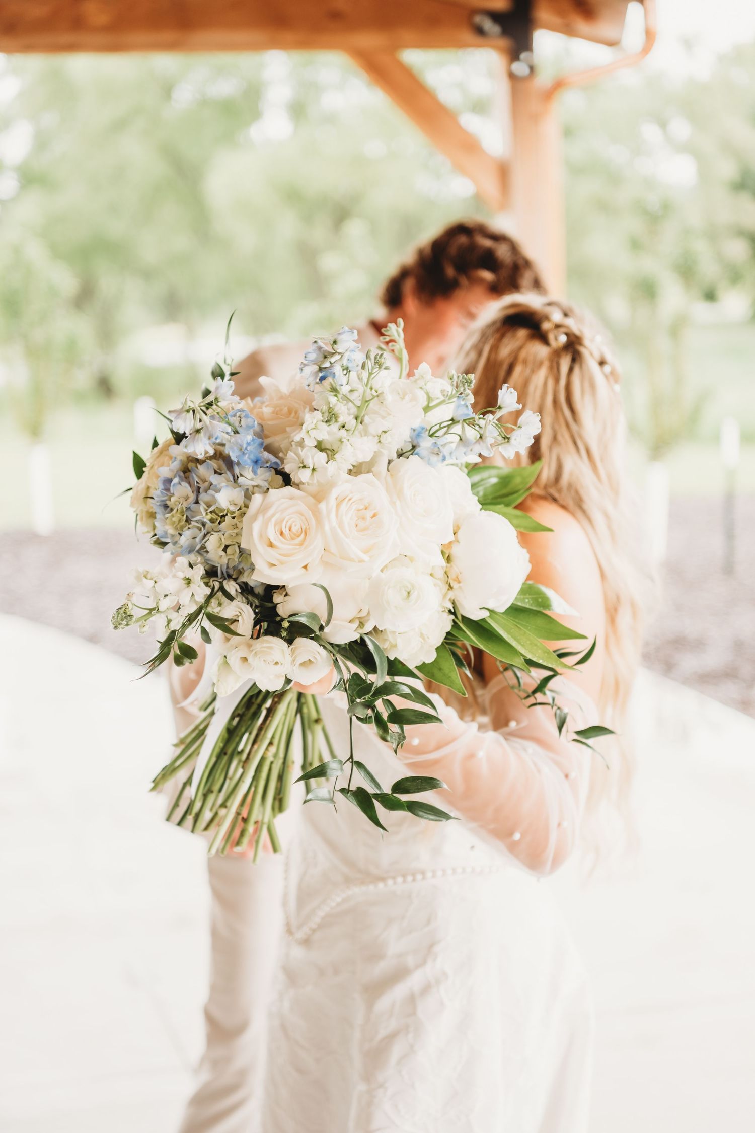 Couple kissing behind a large white and blue bridal bouquet of roses under a wooden pavilion.