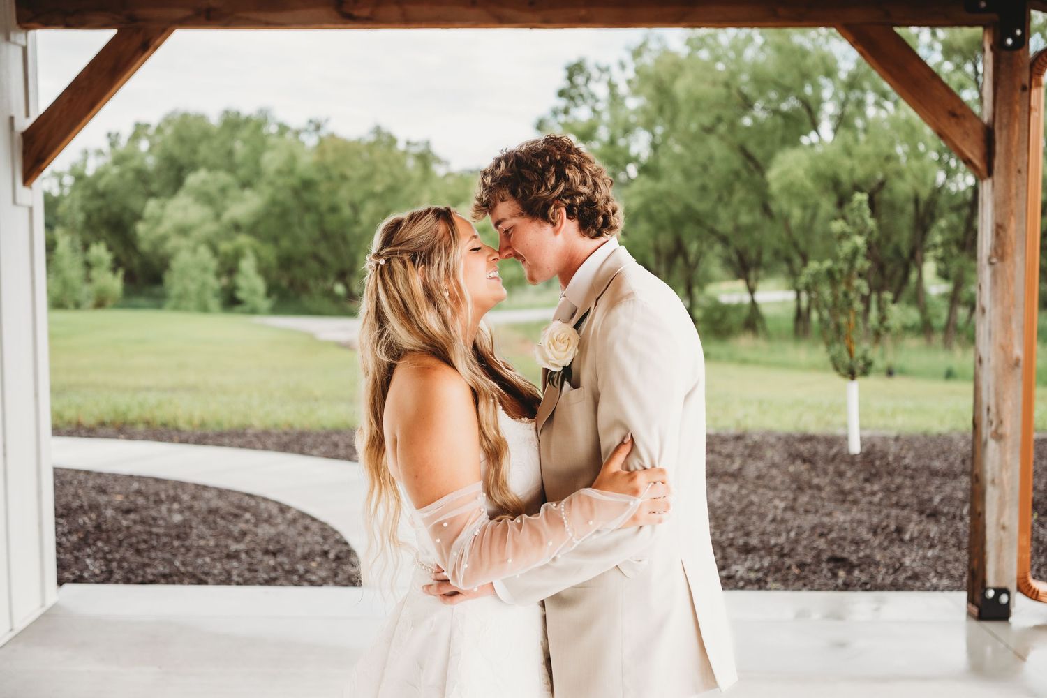 A bride and groom share a romantic moment under a white pavilion surrounded by lush greenery on their wedding day.