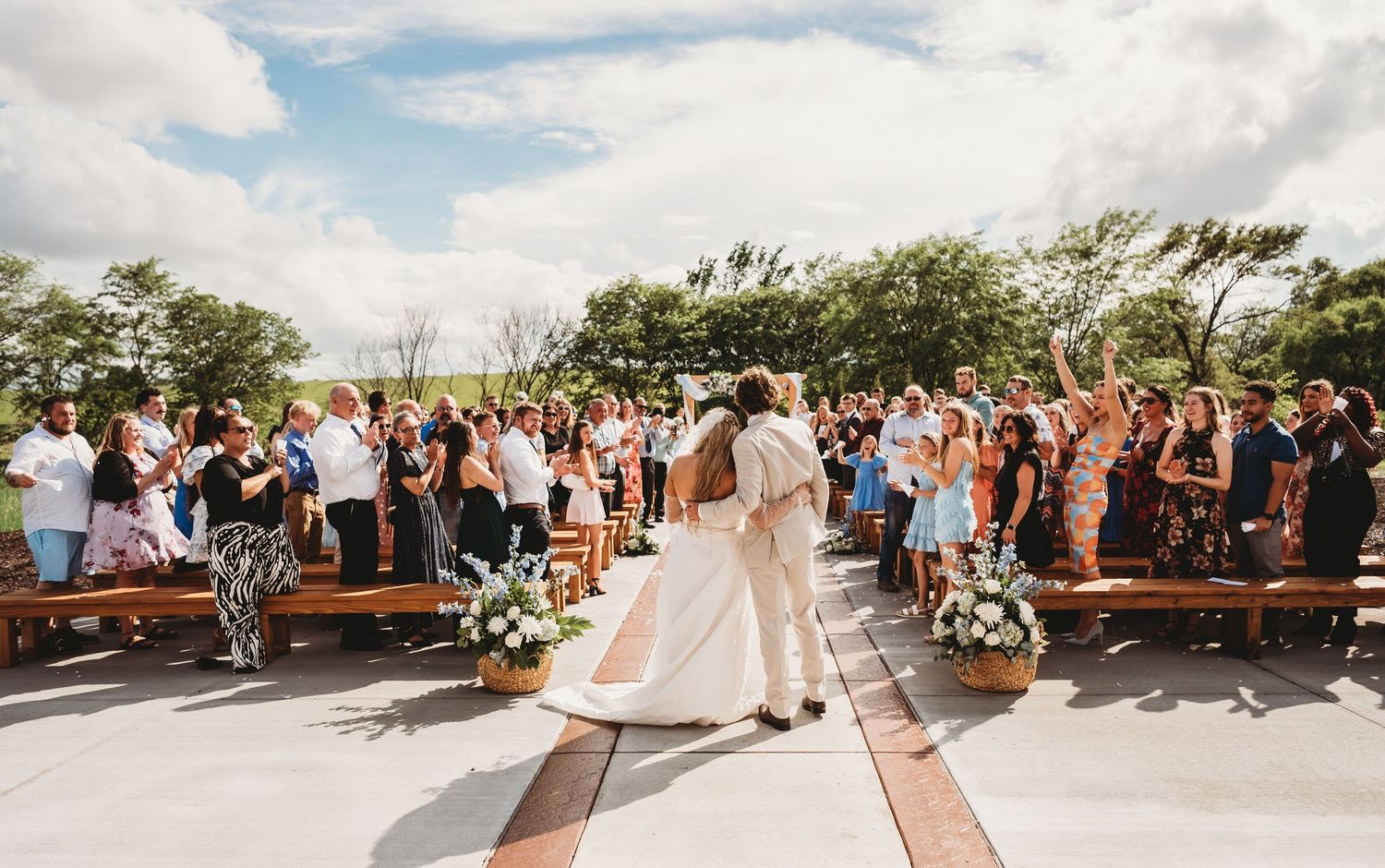 Bride and groom share first kiss at outdoor wedding ceremony with guests cheering on wooden pews.
