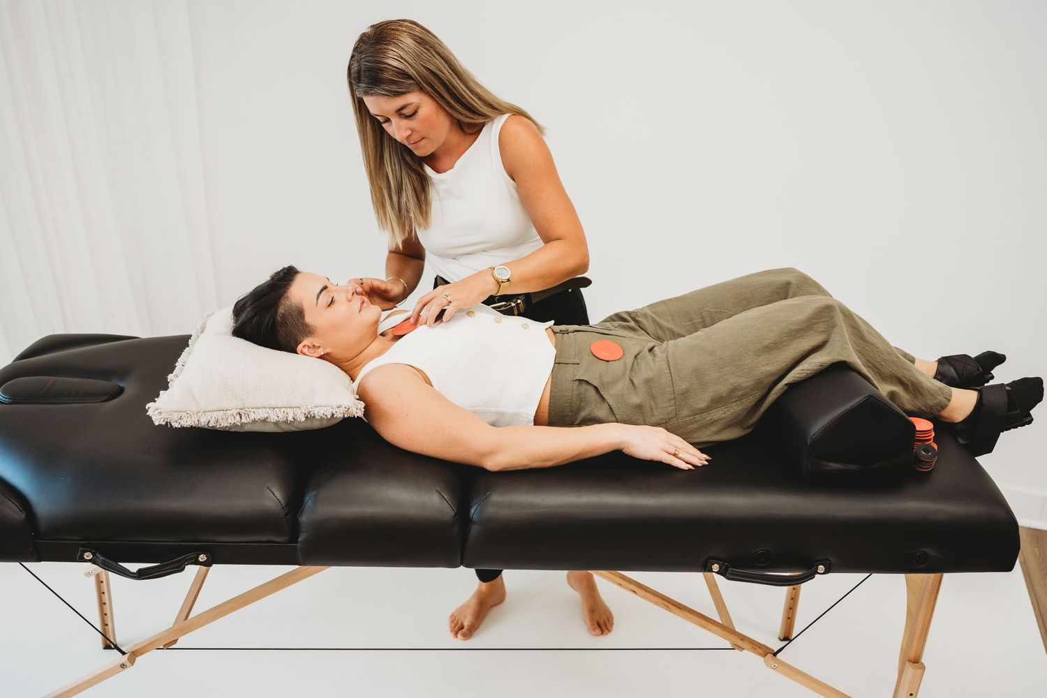 Therapist performing a neck and shoulder massage on a client lying on a black massage table in a bright studio.