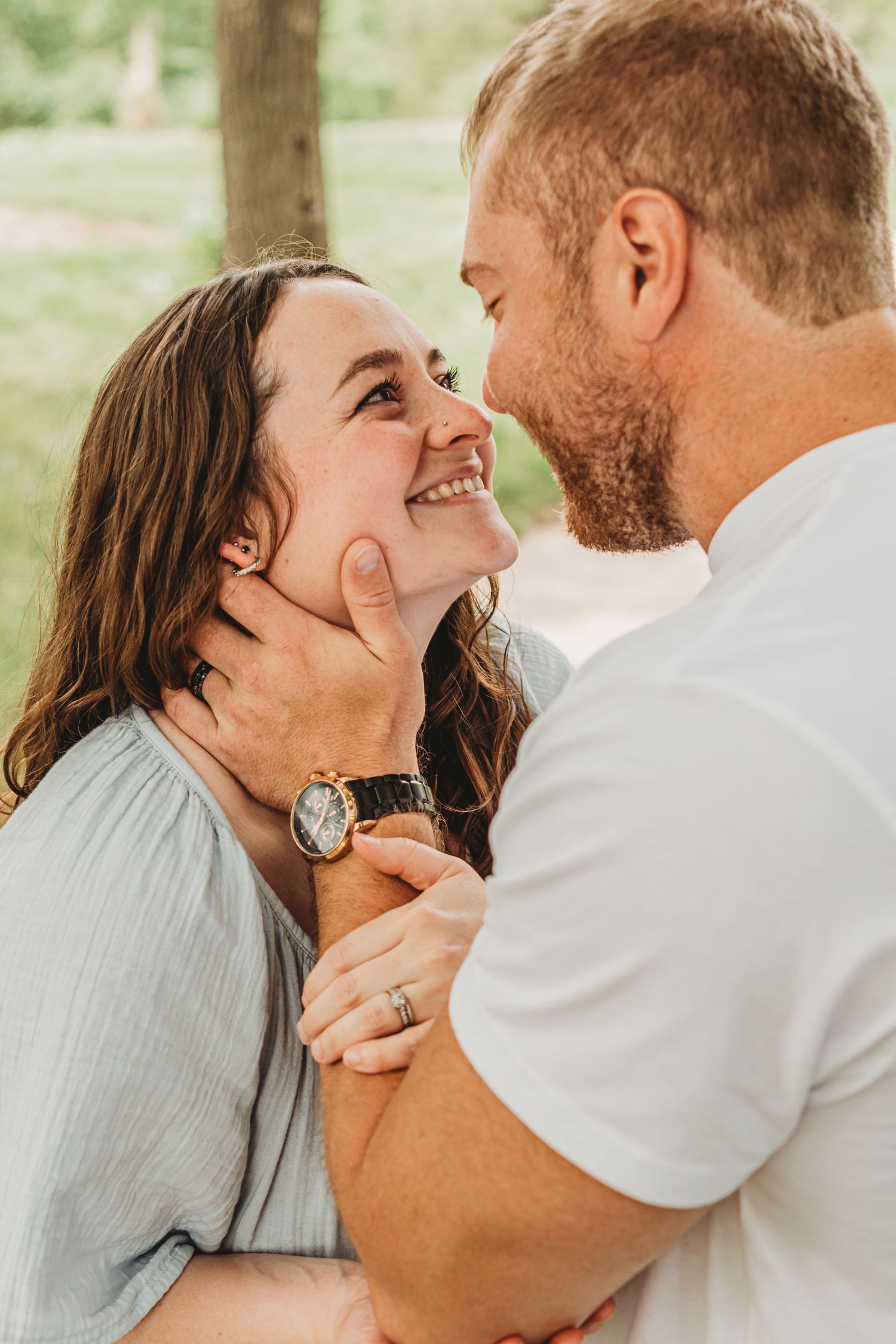 A man tenderly cups a laughing woman's face as they share a close romantic moment outdoors.