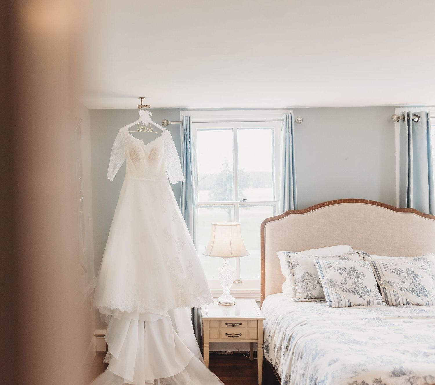 Elegant white wedding dress hanging near a window in a softly decorated bedroom with a pink headboard.