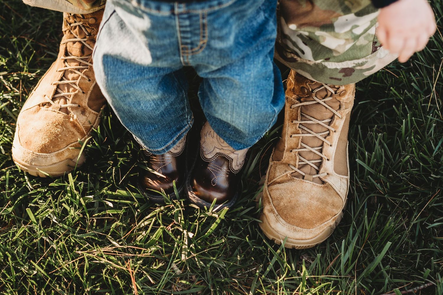 Adult military boots beside small child cowboy boots on green grass.