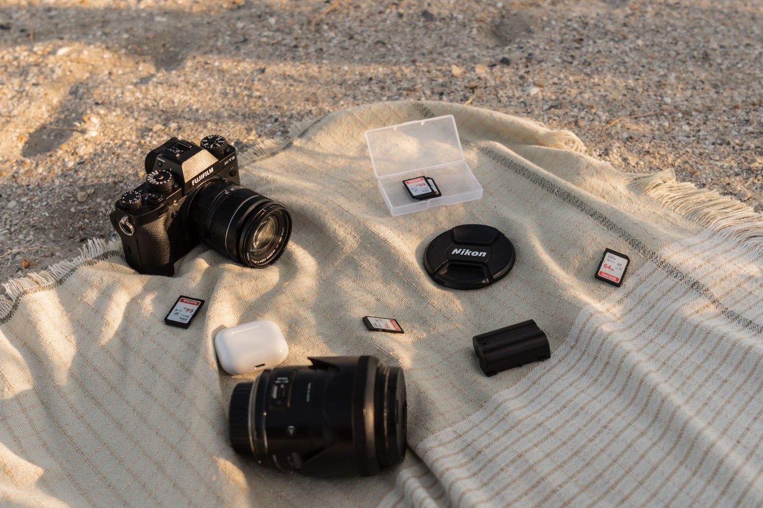 Flat lay of Nikon camera, lenses, SD cards, and battery on a sandy beach blanket.