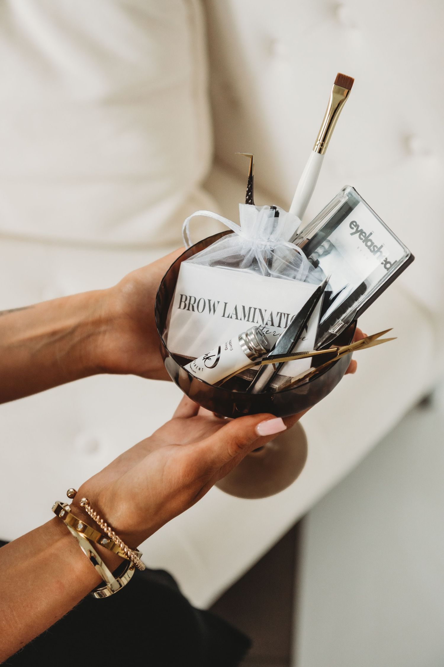 Hands holding a decorative bowl filled with brow lamination beauty tools and brushes.