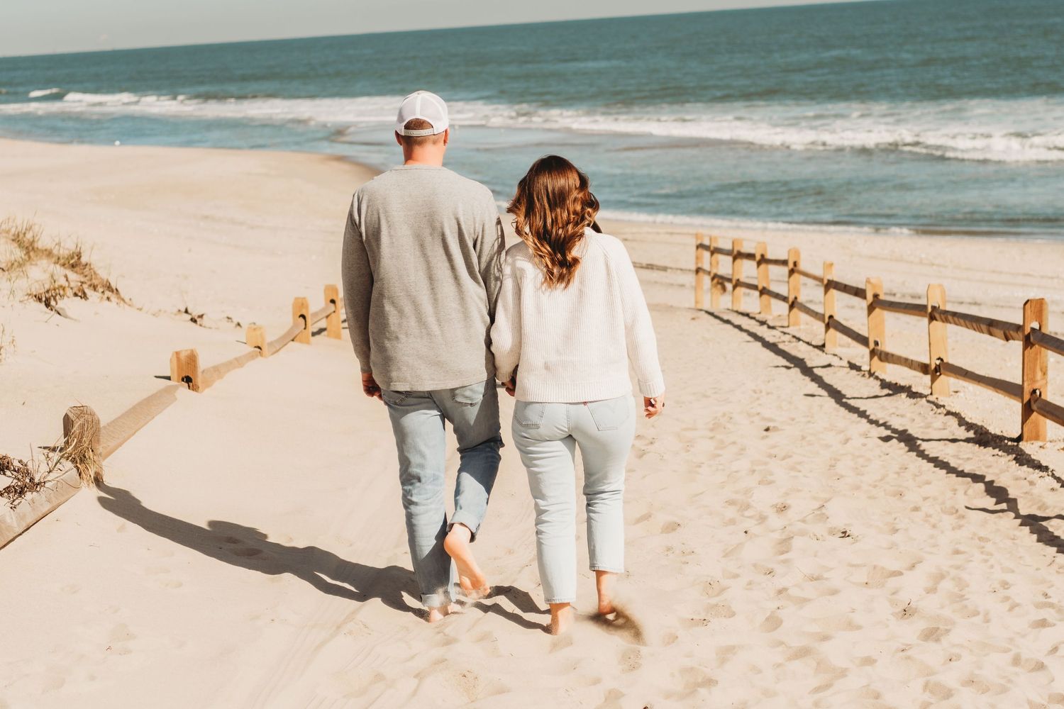 Couple walking hand in hand on sandy beach boardwalk path toward the ocean.